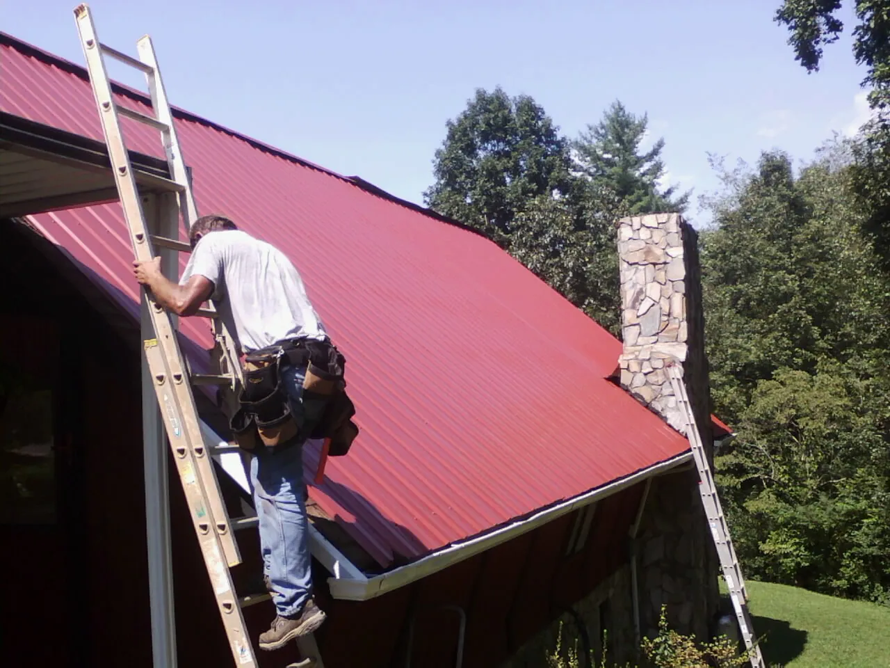 Roofer on a ladder beside a red metal roof and stone chimney — Soffit & Fascia Repair by Roofing Brothers in Onamia
